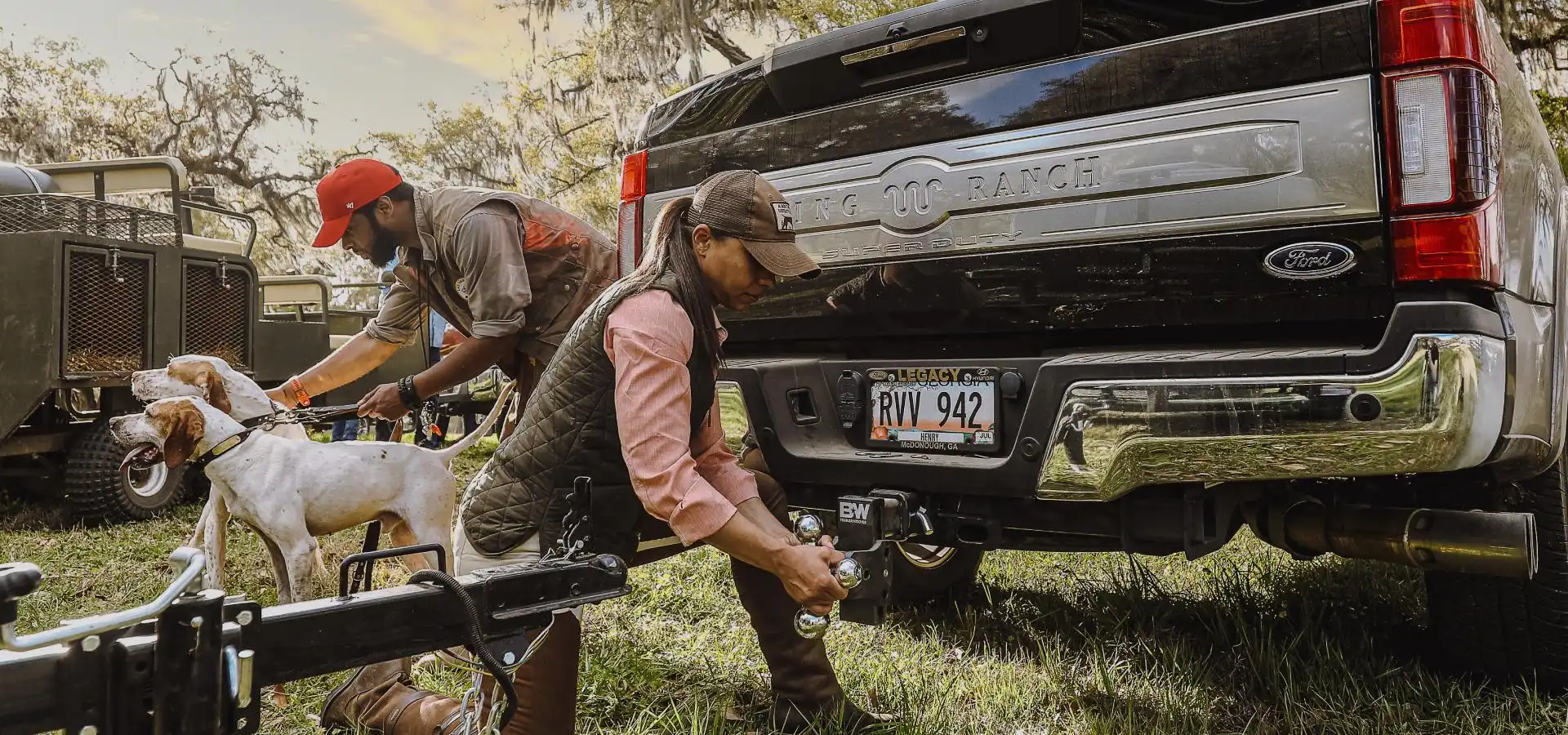 Man and woman with dogs hitching a trailer to a pickup truck with parts from Duke's A&W, preparing to travel in the outdoors